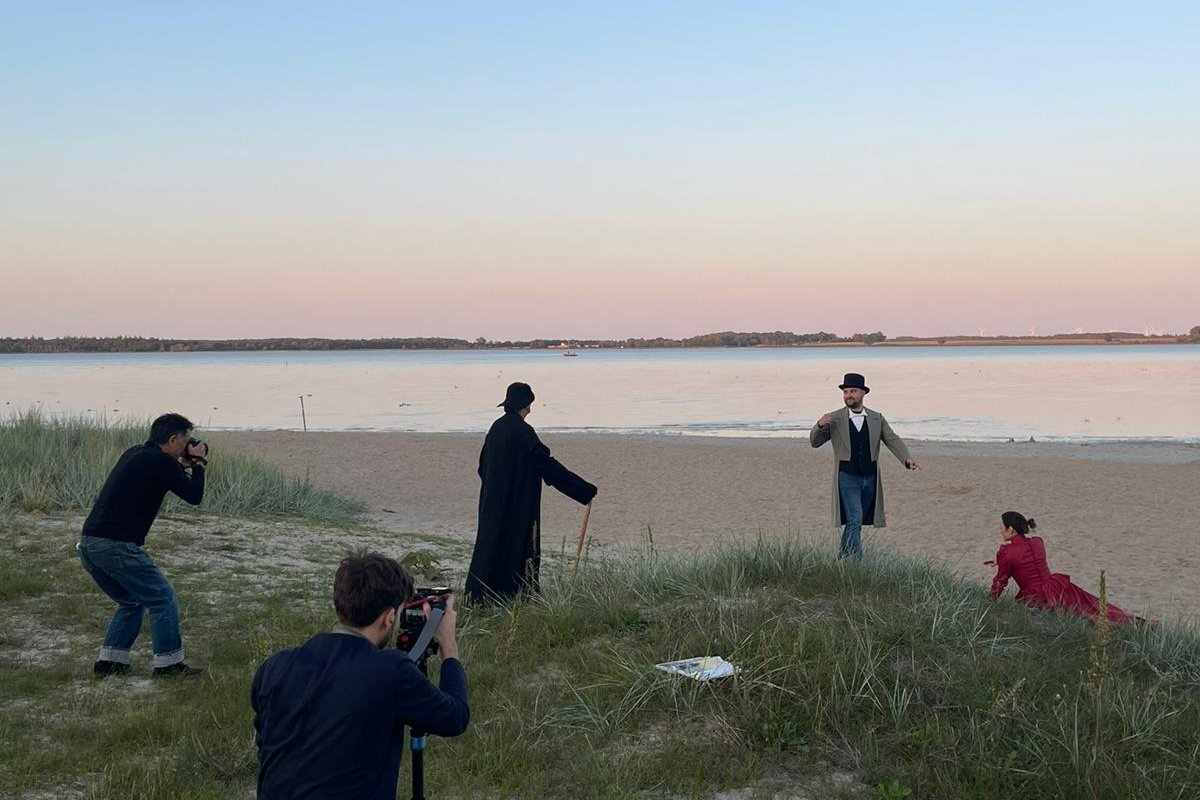 Foto: Obere Bidlhälfte blauer Himmel bei Sonnenuntergang. Untere Bildhälfte Meer, Strand und zum Großteil ein Grasvorsprung, auf dem drei Personen in historischen Kostümen für zwei Fotografen im Vordergrund posieren.