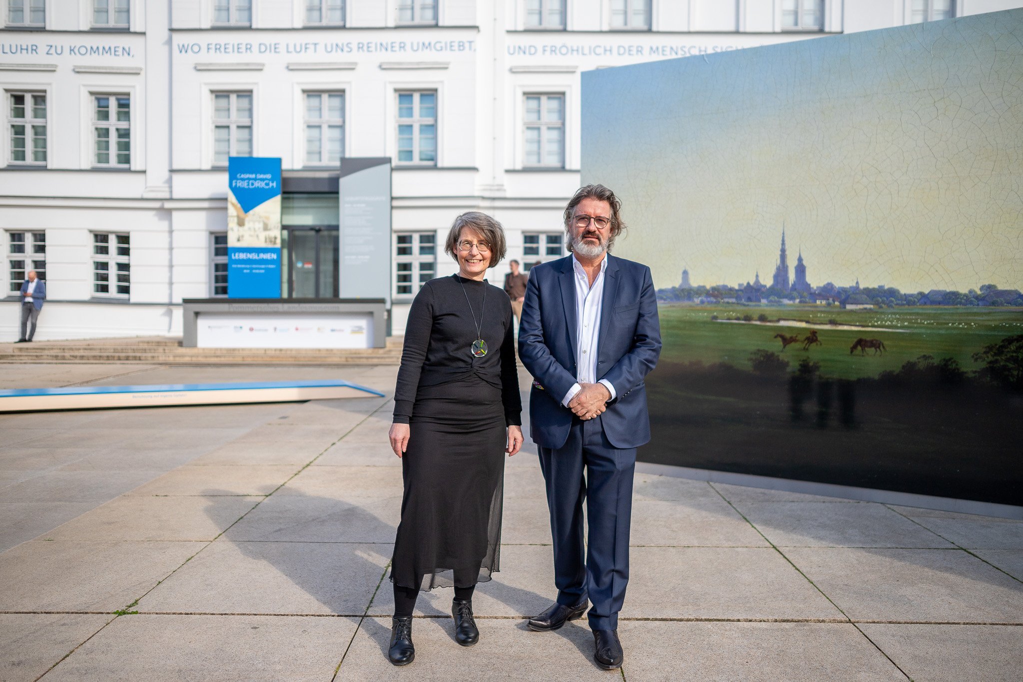 Dr. Birte Frenssen und Olafur Eliasson, hinter ihnen auf dem Vorplatz ein großer Aufsteller mit den "Wiesen bei Greifswald", dahinter die Fassade des Museums.