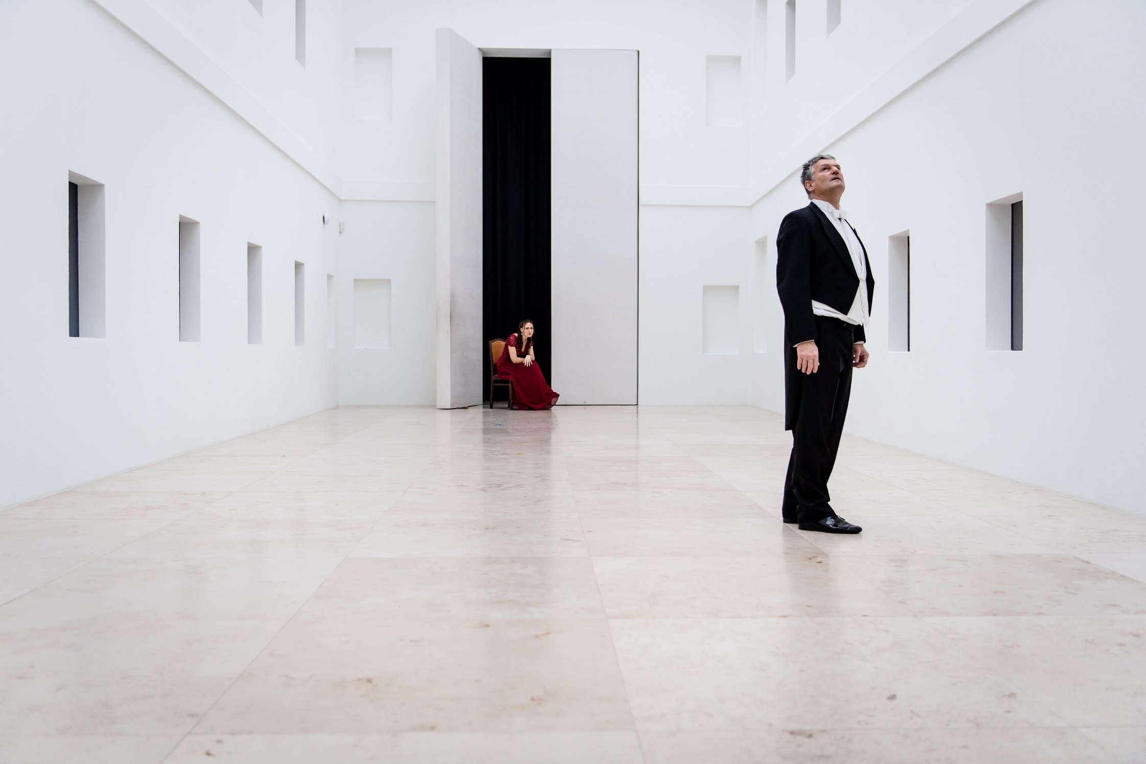 Weißer Innenhof des Pommerschen Landesmuseums. In der Tür im Hintergrund sitzt eine Frau in rotem Kleid auf einem Stuhl. Im Vordergrund rechts ein stehender Herr im Frack, welcher nach oben blickt.