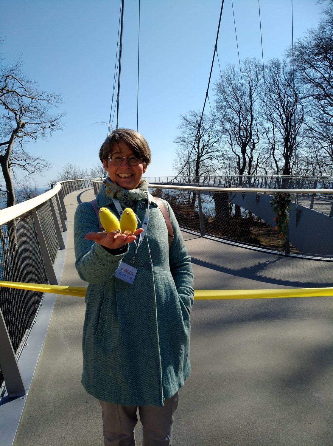 Dr. Birte Frenssen steht vor der Plattform des neuen Skywalks, noch mit einem gelben Ban abgesperrt. In ihrer rechten Hand präsentiert sie die Kanarienvögel Roman & Tik. Im Hintergrund strahlend blauer Himmel.