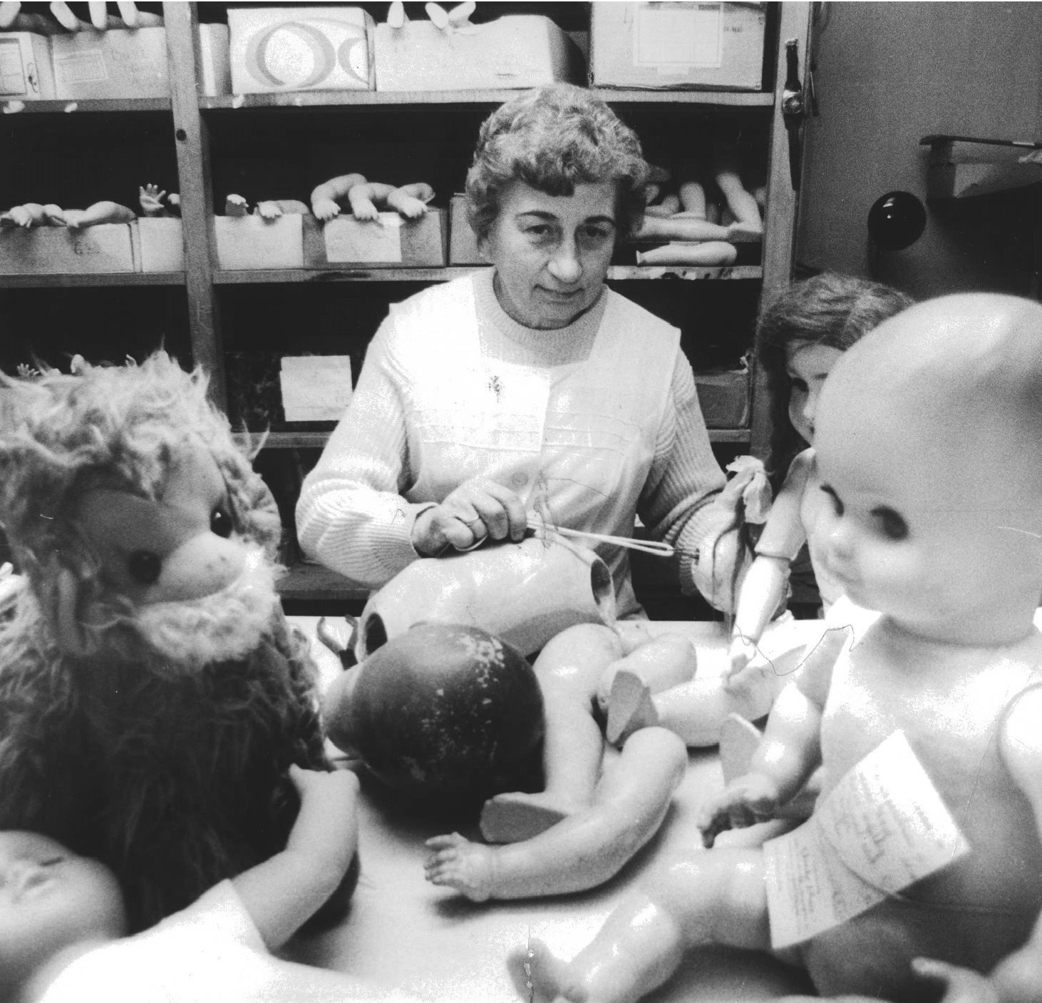 Die Greifswalder Puppendoktorin Gerda Baer bei der Arbeit: Das Schwarz-Weiß-Foto zeigt sie inmitten von Puppen, Puppenkörperteilen und Stofftieren.
