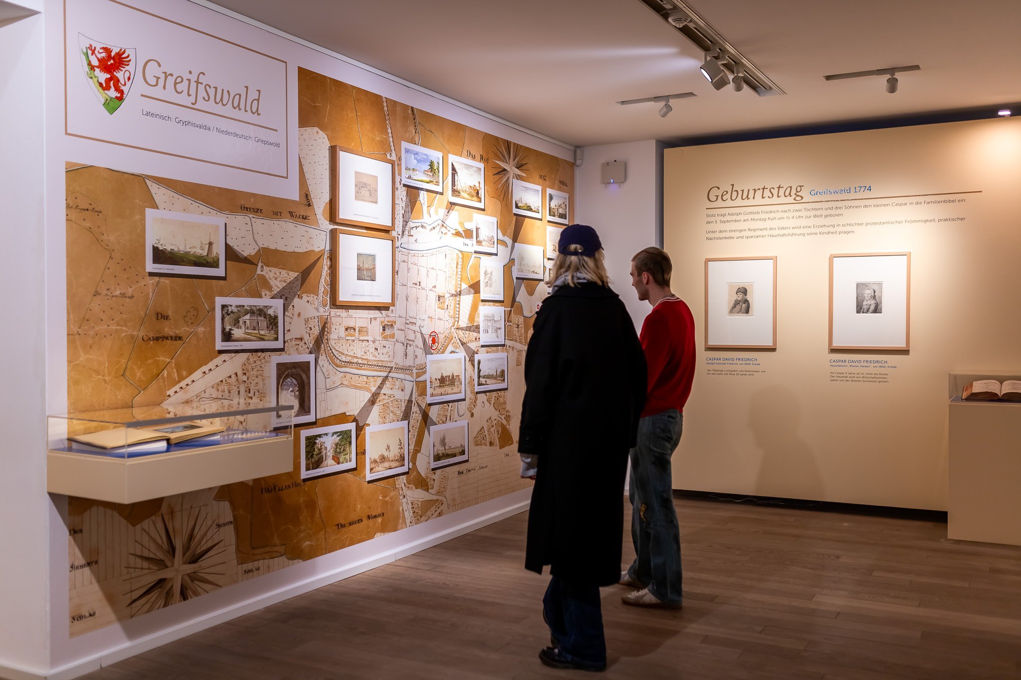 2 Besucher*innen in der Ausstellung, an der linken Wand eine große Karte, rechts zwei Grafiken.