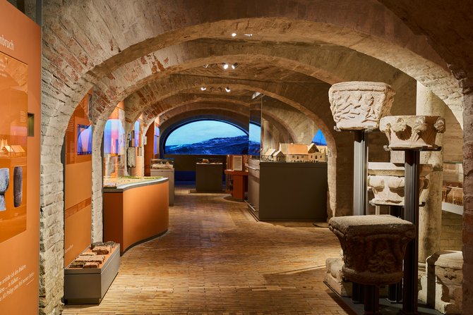 Blick in die Ausstellung: das Mittelalter, die Zeit der Hanse - Ausstellungsraum mit hellem Backsteingewölbe, orangenen Ausstellungsflächen sowie ausgestellte Exponate