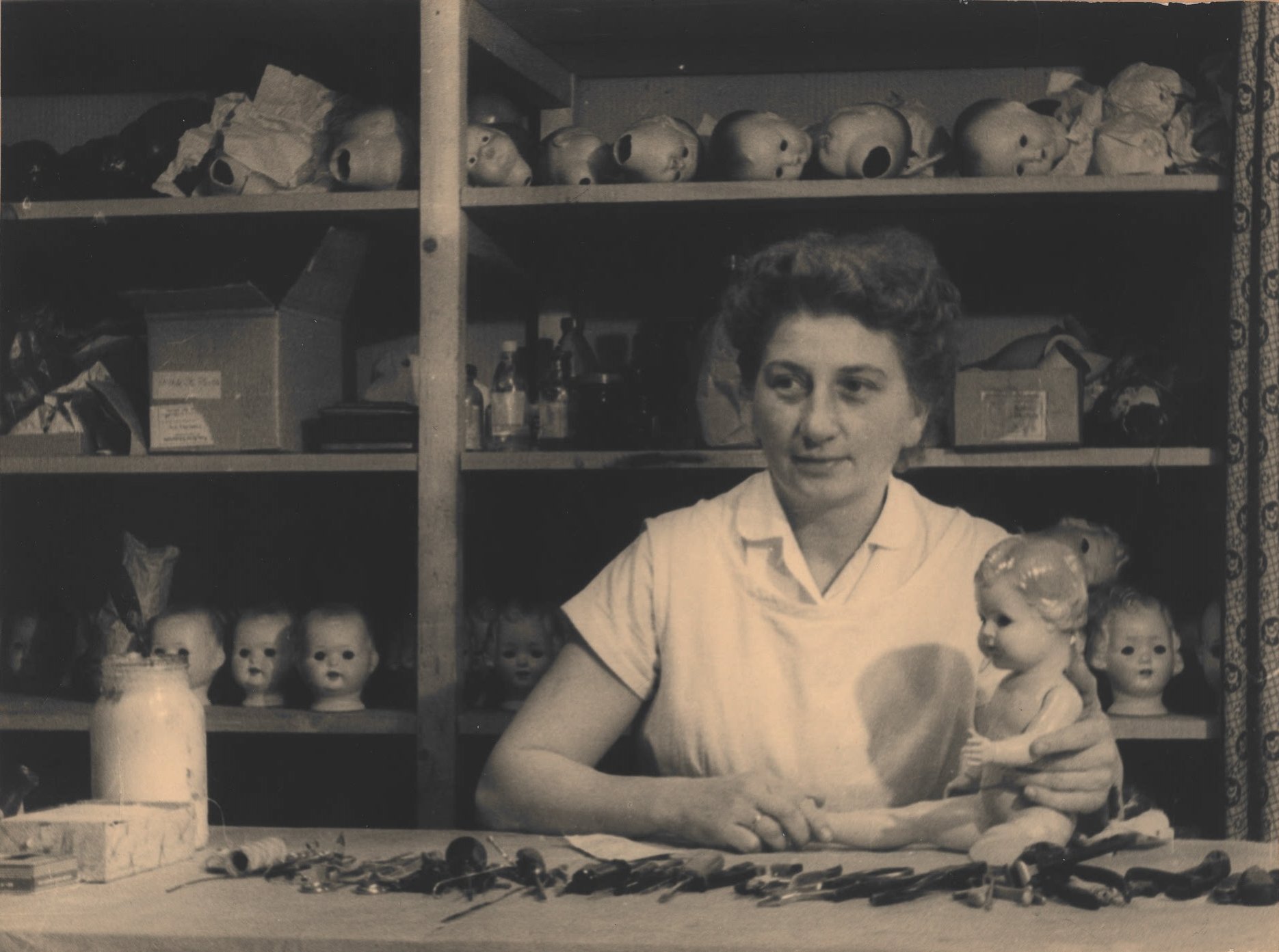 Die Greifswalder Puppendoktorin Gerda Baer bei der Arbeit: Das Schwarz-Weiß-Foto zeigt an einem Tisch sitzend, mit einer Puppe und vielen Werkzeugen. Hinter ihr ein großes Regal mit vielen Puppenköpfen, Kartons, Flaschen.