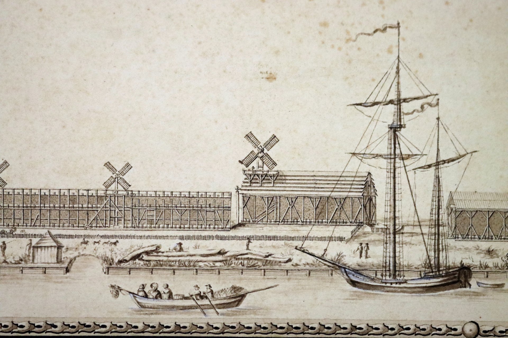 Schwarz-weiß Grafik. Im Vordergrund ein Fluss mit Ruderboot und Segelboot, dahinter Gebäude aus Holzbalken, darauf Windmühlen.