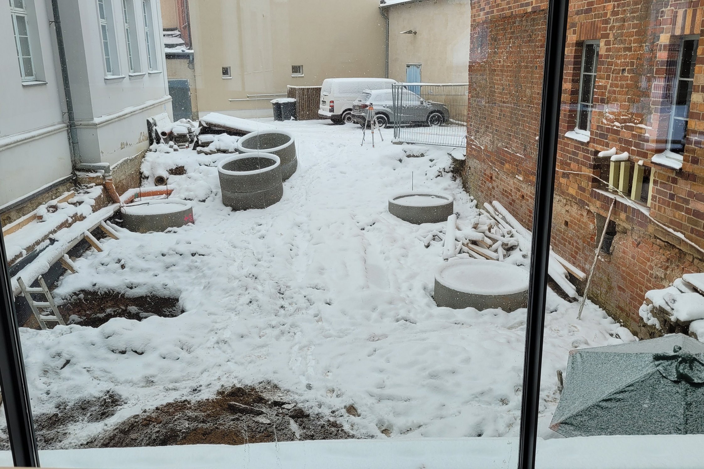 Blick auf das Areal der Ausgrabung von der Museumsstraße aus, rechts und links Gebäude. Auf der Erdoberfläche liegt Schnee. Zu erkennen sind Löcher, teils mit Brunnenringen, in zwei Reihen.