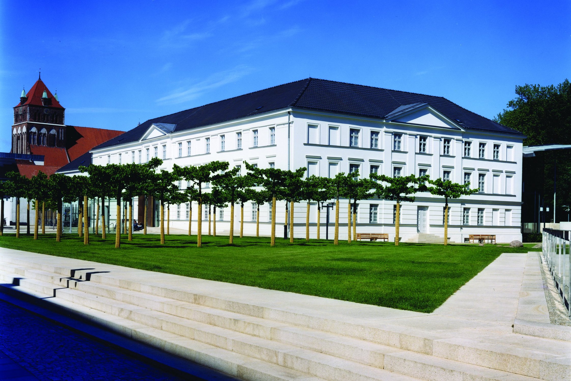 Das Pommersche Landesmuseum inmitten seiner Gärten: Der weiße Bau des Grauen Klosters liegt hinter Bäumen und einem Rasen mit Findlingen 