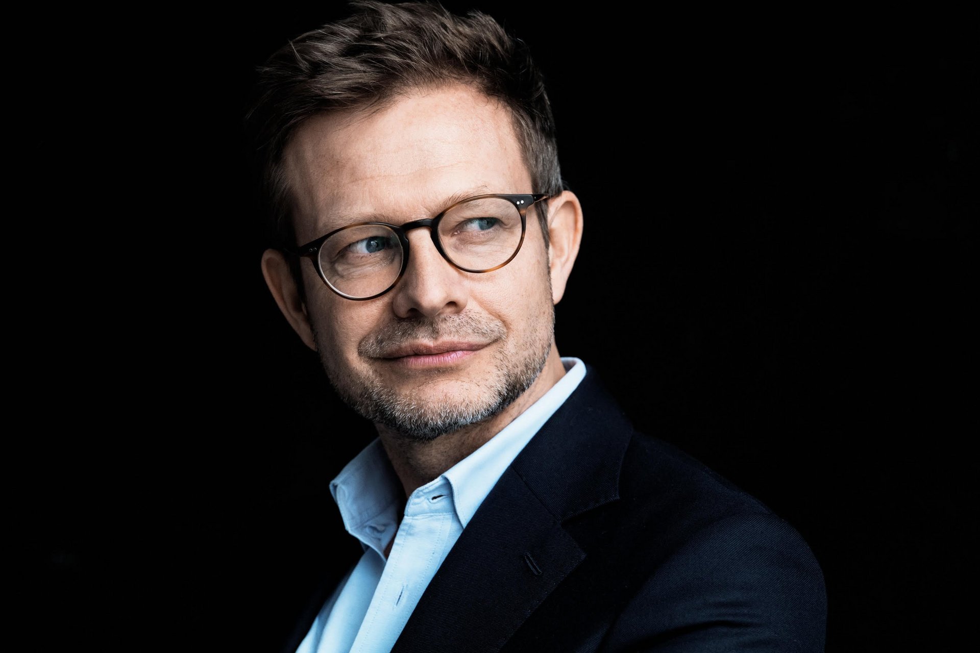 Portraitfoto: Mann mit Brille und dunkelblauem Anzug nach links gewendet, schaut nach rechts vor schwarzem Hintergrund.