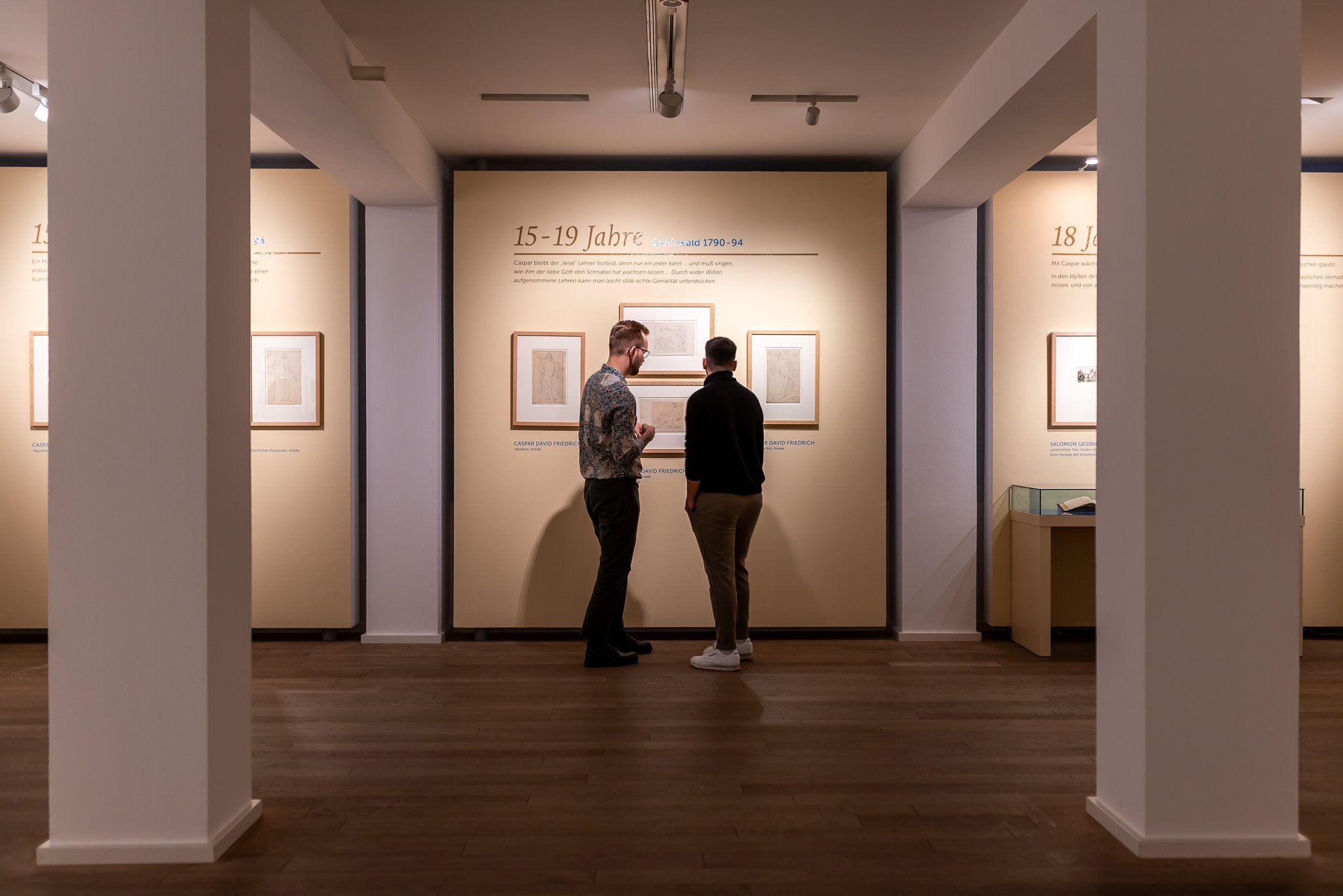 Blick in den Ausstellungsraum. Zwischen zwei Pfeilern hinten zwei Besucher, die mehrere Grafiken betrachten.
